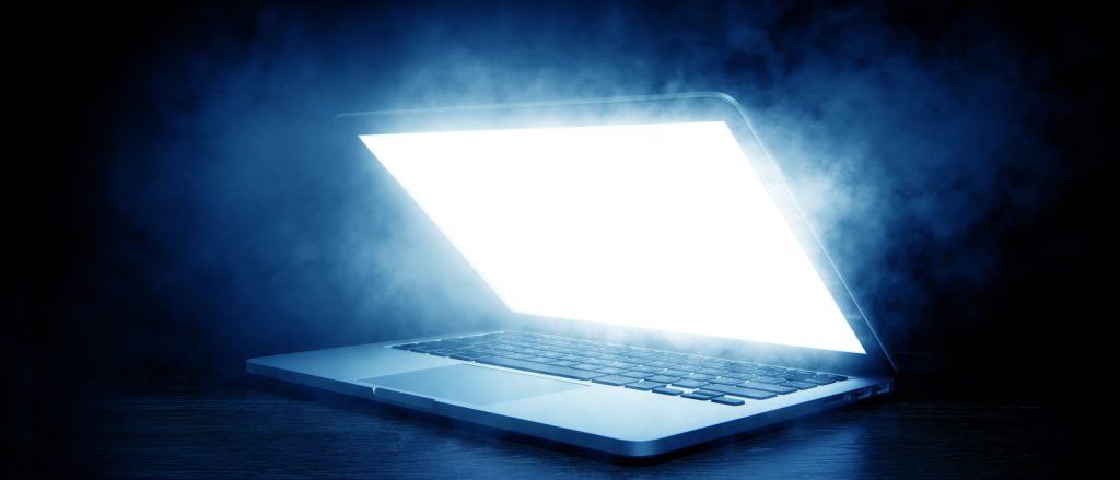 A laptop emits a bright white glow in a dark setting, surrounded by mist