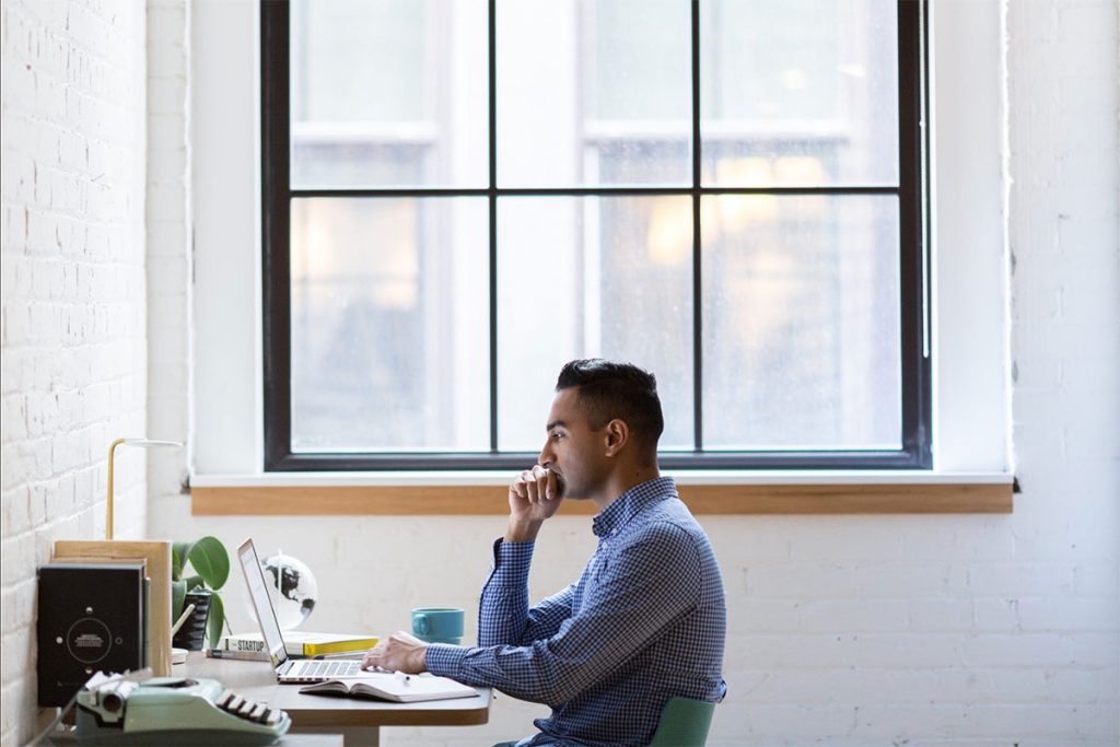 A person sits at a desk in front of a large window, working on a laptop and resting their chin on their hand