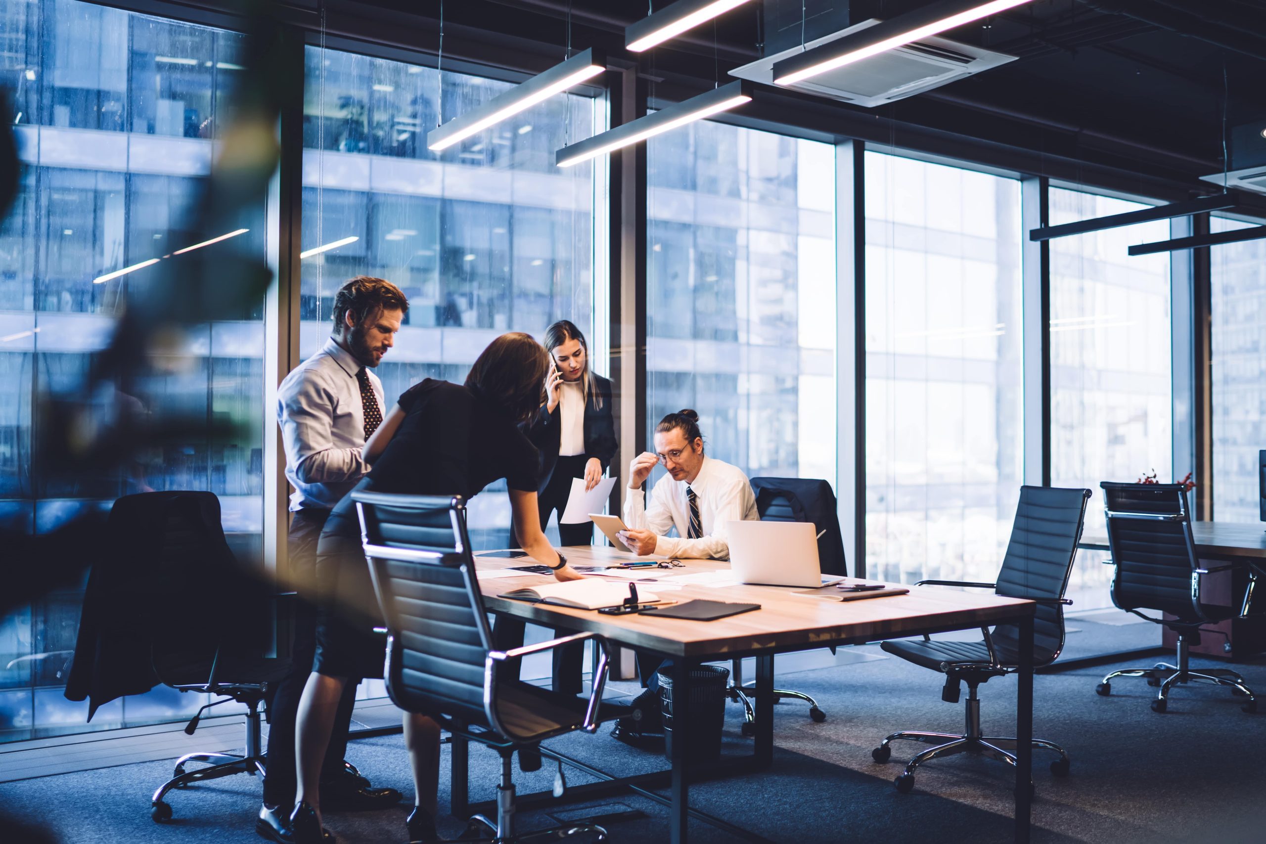 Four business professionals collaborate around a table in a modern office with large windows, glass walls, and city views. Laptops and documents are on the table, and sunlight brightens the workspace.